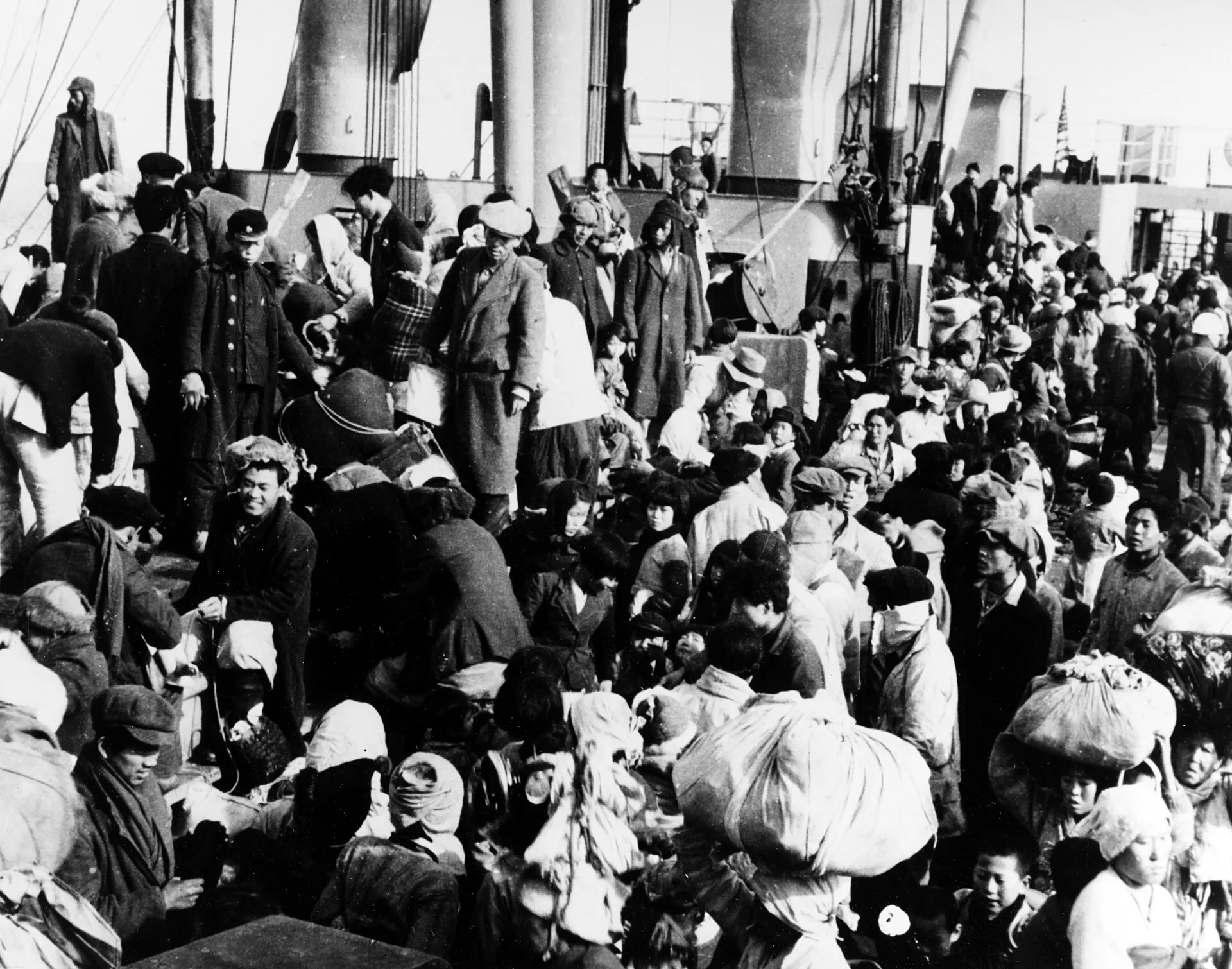 Hundreds of Korean refugees packed shoulder to shoulder with their bundled belongings on the deck of a ship, an American flag visible above them