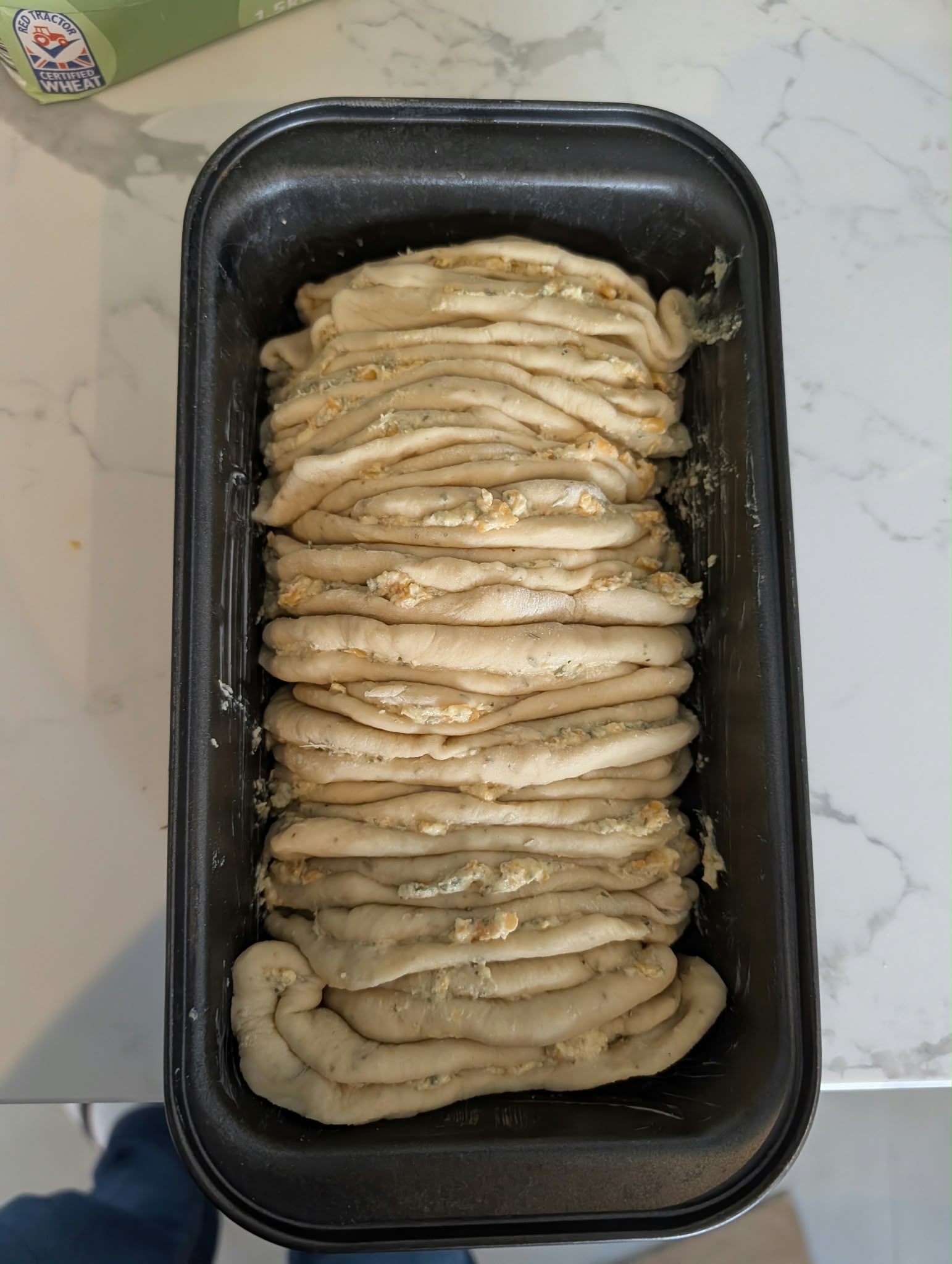 Twelve folded rounds of dough lined up on edge in a black loaf pan, herb butter and shredded cheese visible in every seam